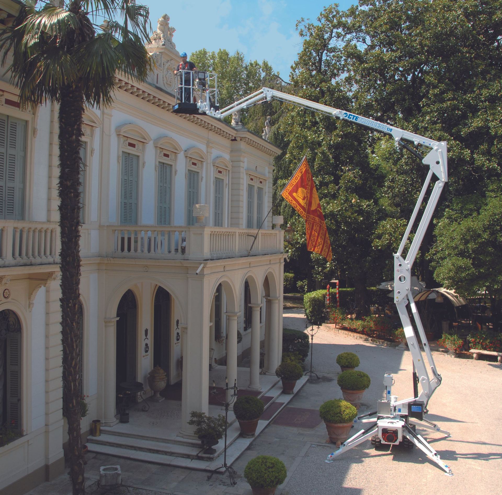 Person on a lift working on the roof of a large, white building. Lift is extended; a garden surrounds.