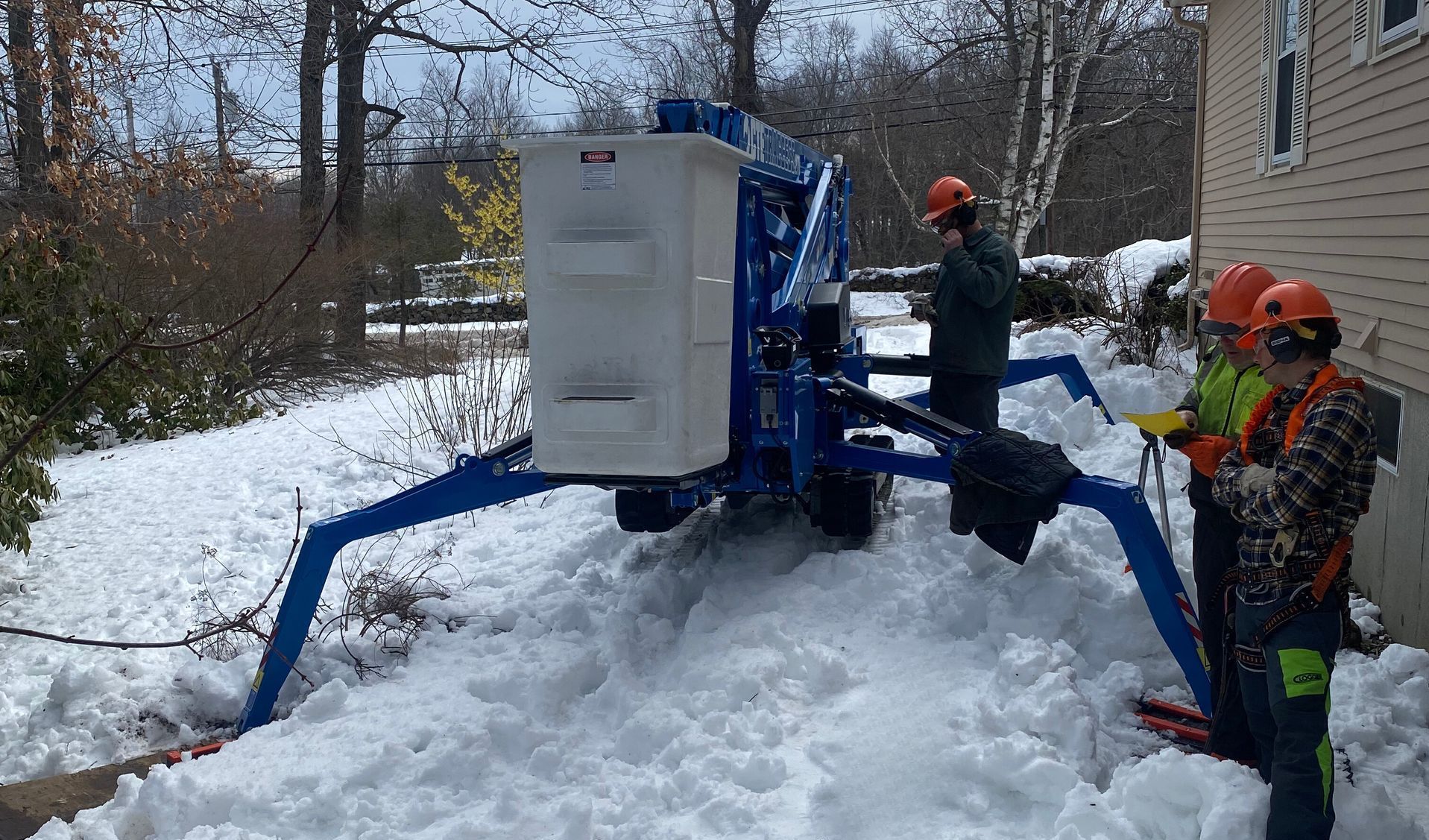 Blue lift with three people in helmets, near a house, in snow.