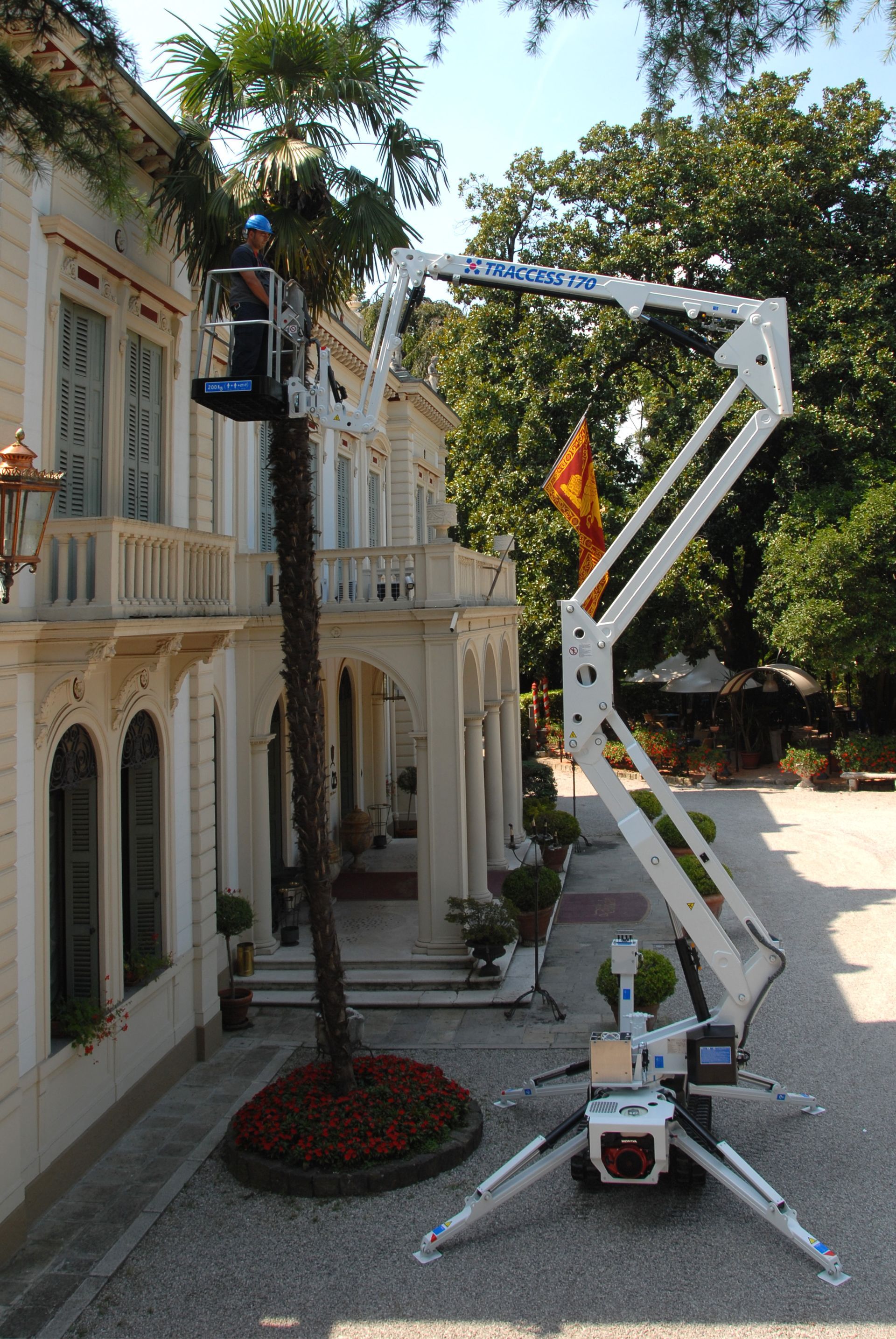Man in lift trimming a tree near a white building with columns and a balcony. White lift, sunny day.