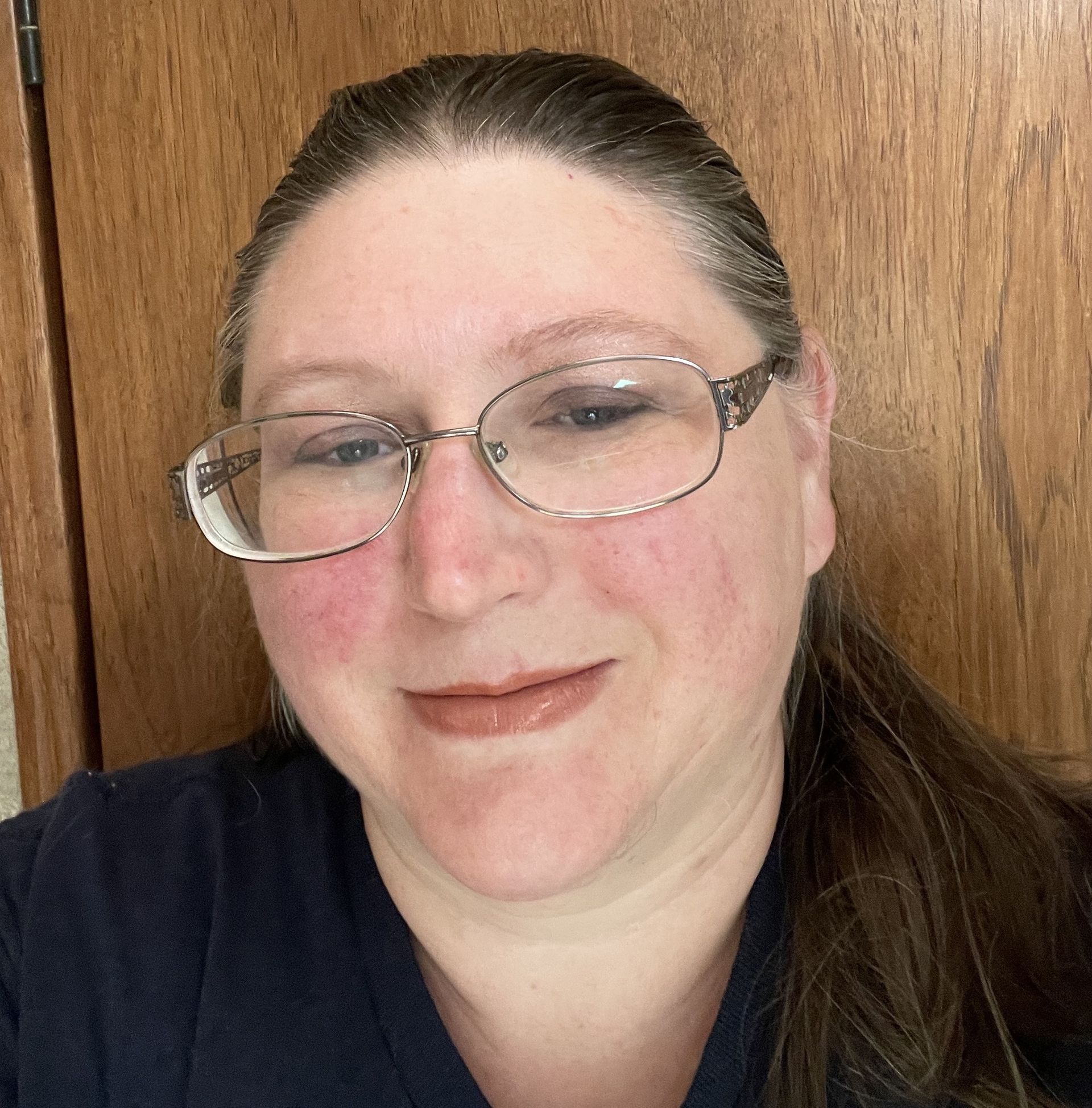 A woman wearing glasses is smiling in front of a wooden door.