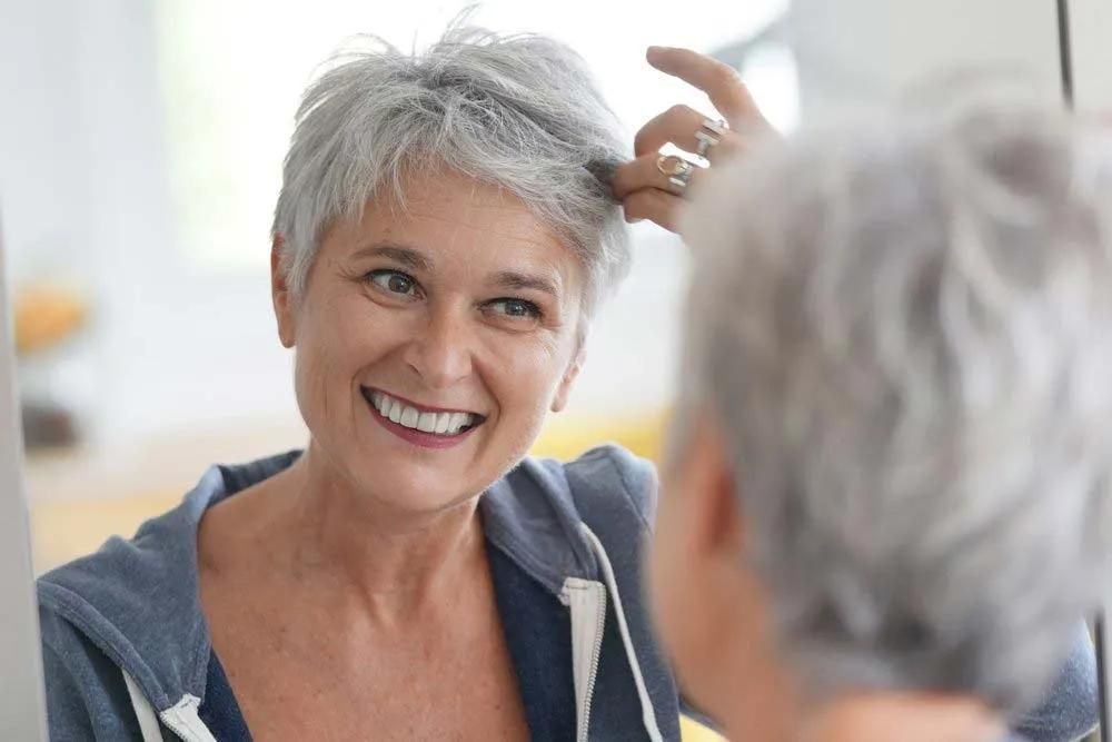 A Woman is Brushing Her Gray Hair in Front of a Mirror — Southside Hair & Beauty Design In South Tamworth, NSW A Woman is Brushing Her Gray Hair in Front of a Mirror — Southside Hair & Beauty Design In South Tamworth, NSW