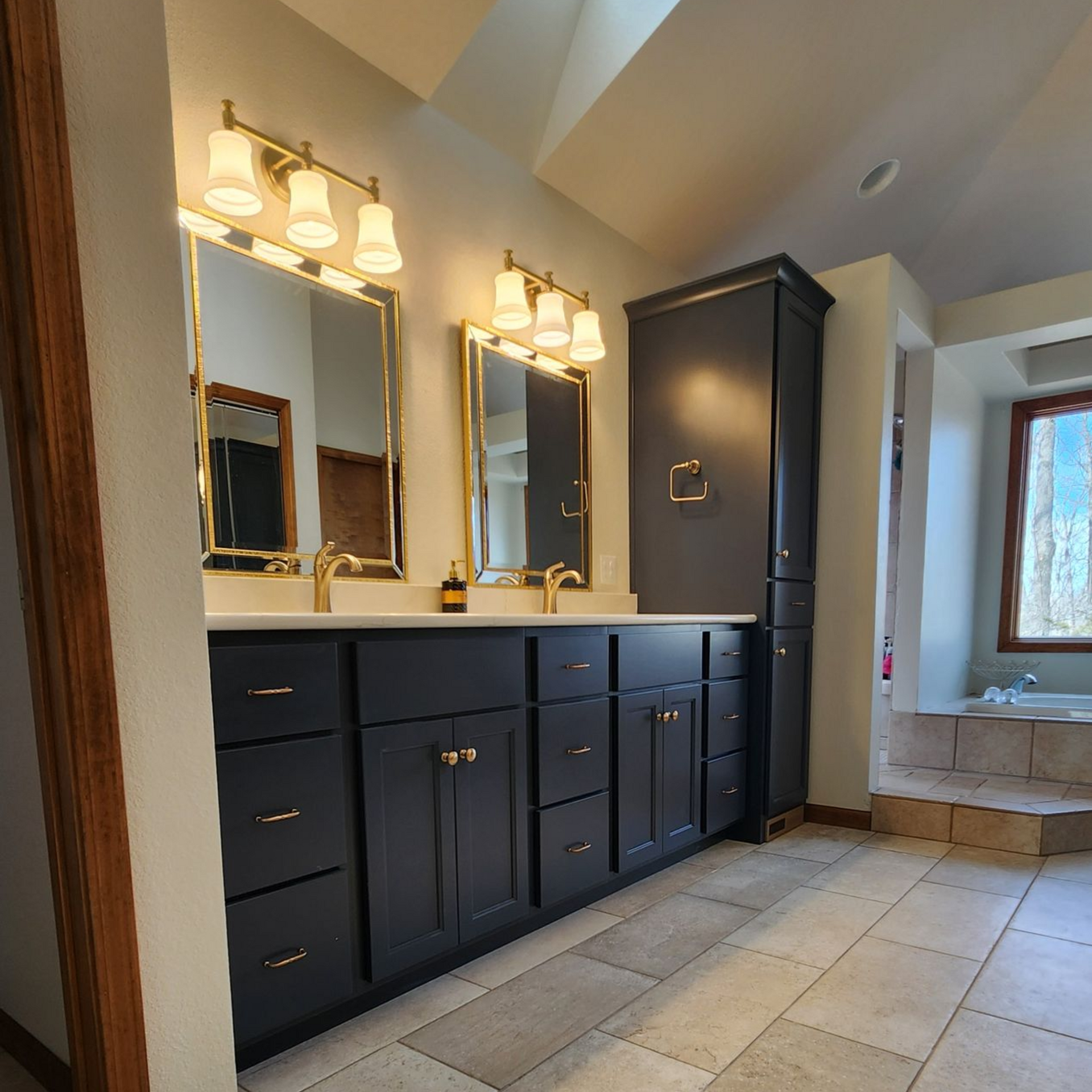 Bathroom with navy blue vanity, gold fixtures, mirrors, and storage cabinet. Beige tile flooring and natural light.