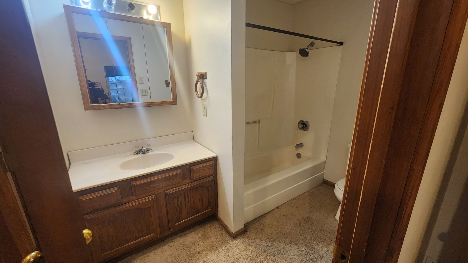 Bathroom with a vanity, mirror, and shower/tub combination. Brown cabinets and tan carpet.