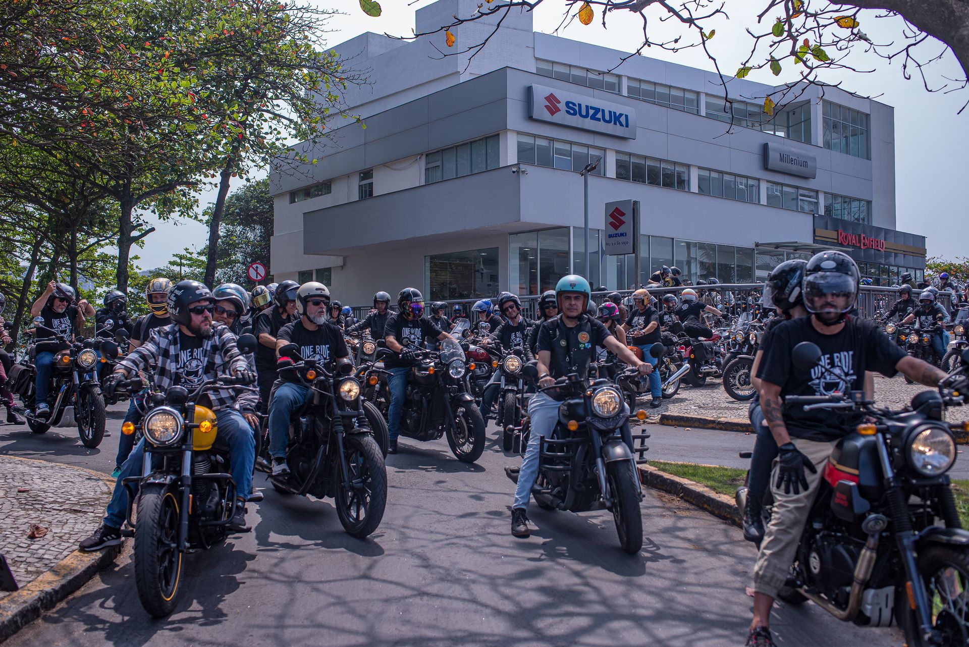 Um grande grupo de motociclistas se reuniu com suas motos em frente a um prédio, sob árvores.
