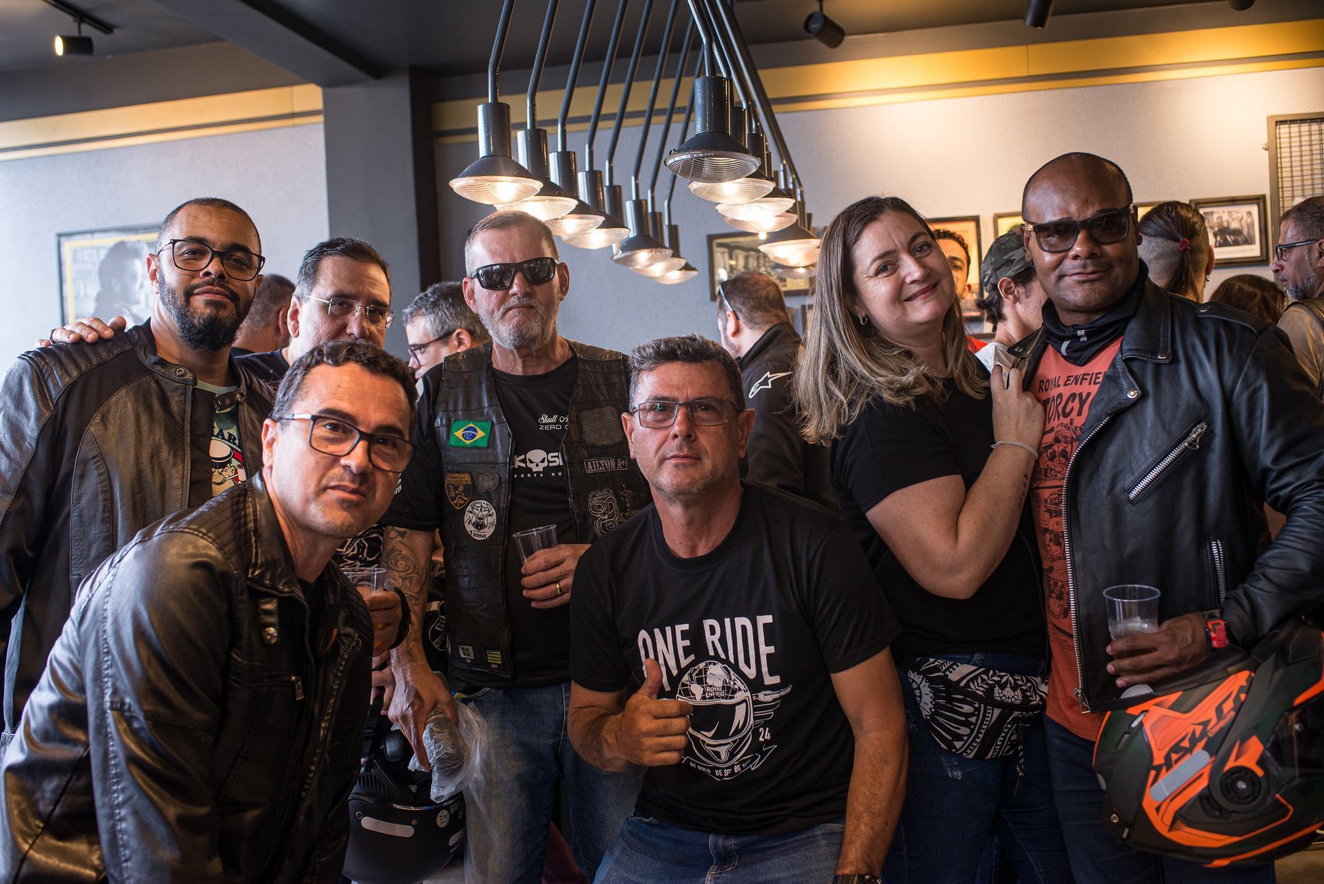 Grupo de seis amigos posando em um bar, sorrindo e segurando bebidas sob luzes pendentes.