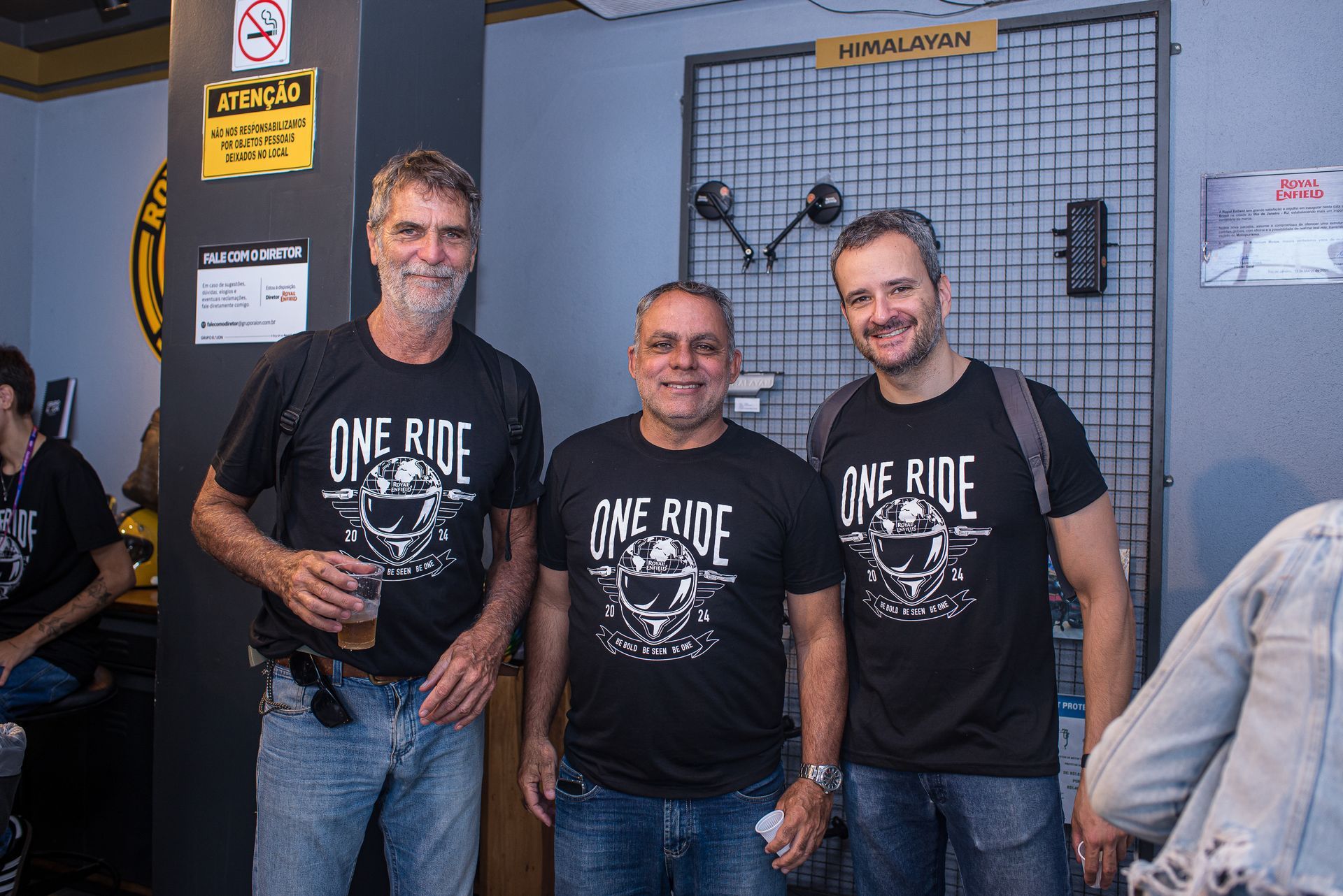 Três homens com camisetas pretas da inscrição “One Ride” posam em um ambiente interno, segurando bebidas e sorrindo.