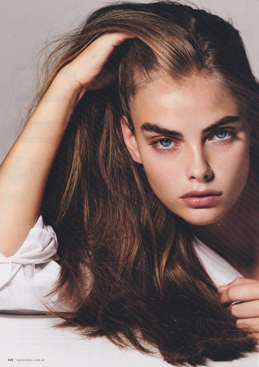 Woman with long brown hair, blue eyes, and full eyebrows. Leaning with hand in hair, wearing white shirt.