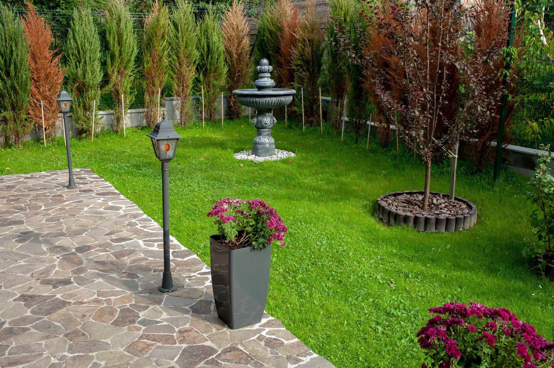 Stone patio beside a green garden with a fountain, lamp posts, and potted flowers