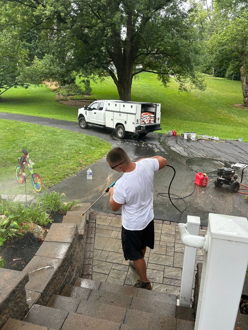Person pressure-washing stone steps beside a yard, with a white van and green trees in the background