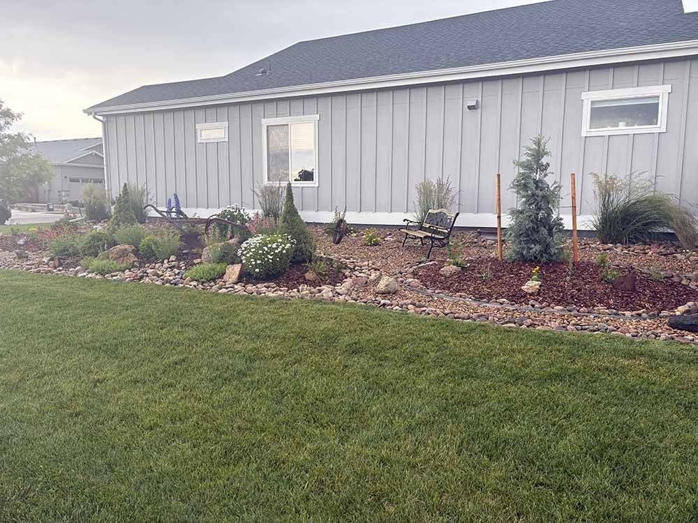 A light gray house with vertical siding and a rock-landscaped yard featuring shrubs and a small bench.