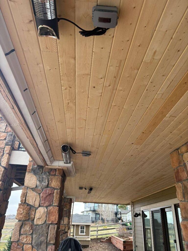 A wooden porch ceiling featuring an outdoor heater, electrical components, and rustic stone pillars under a cloudy sky.