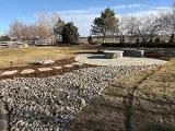 A stone fire pit on a circular stone patio, with a rock riverbed and stone stepping stones leading to it in a backyard.