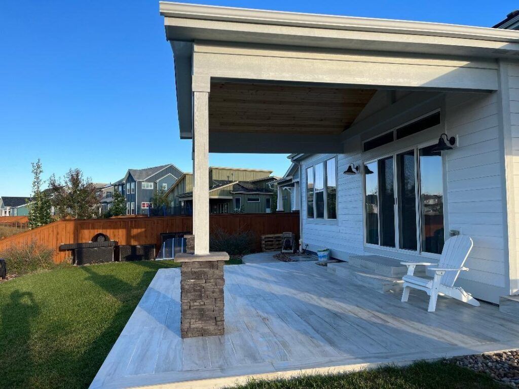 White house patio with stone column support, wood ceiling, light-colored pavers, and a white Adirondack chair.