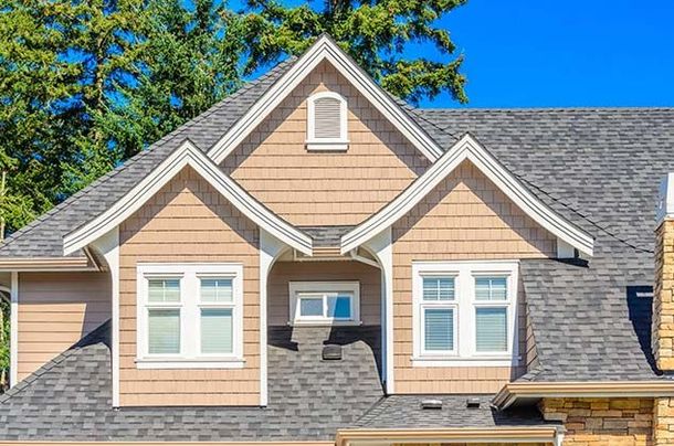 The tan upper floor of a house with two gables, white-framed windows, and grey shingles under a bright blue sky.