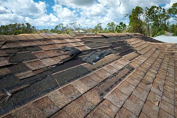 Damaged roof with missing asphalt shingles revealing the dark underlayment against a blue sky with clouds.