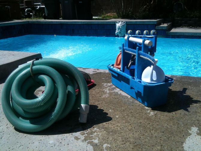 Pool cleaning equipment beside a swimming pool: coiled green hose and blue cart on wet concrete.