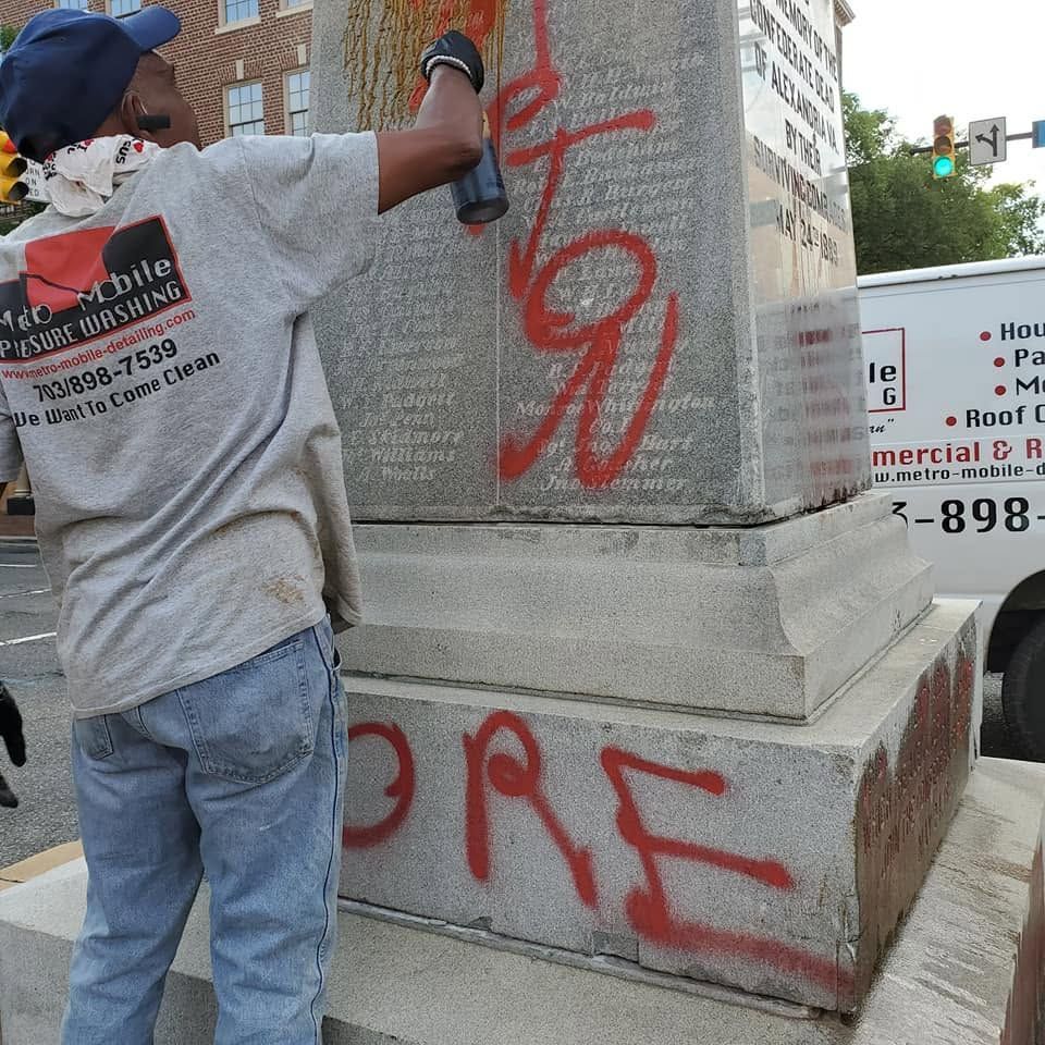 Person spray-painting red graffiti on a stone pillar beside a city street.