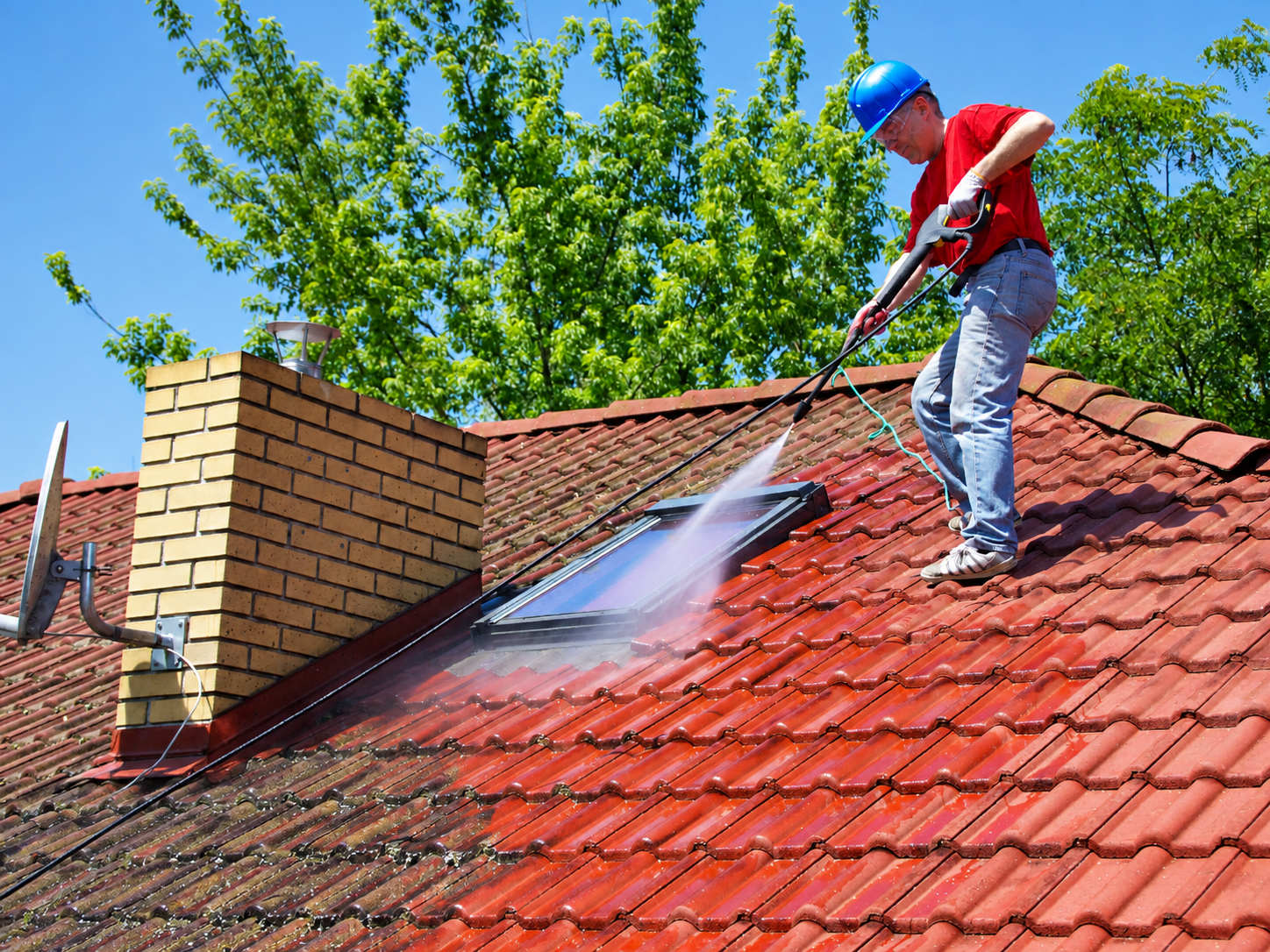 Worker pressure-washing a red tiled roof beside a brick chimney under a blue sky