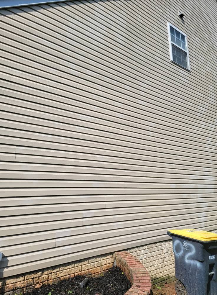 Beige vinyl-sided house wall with a small window, brick planter bed, and trash bins at the edge