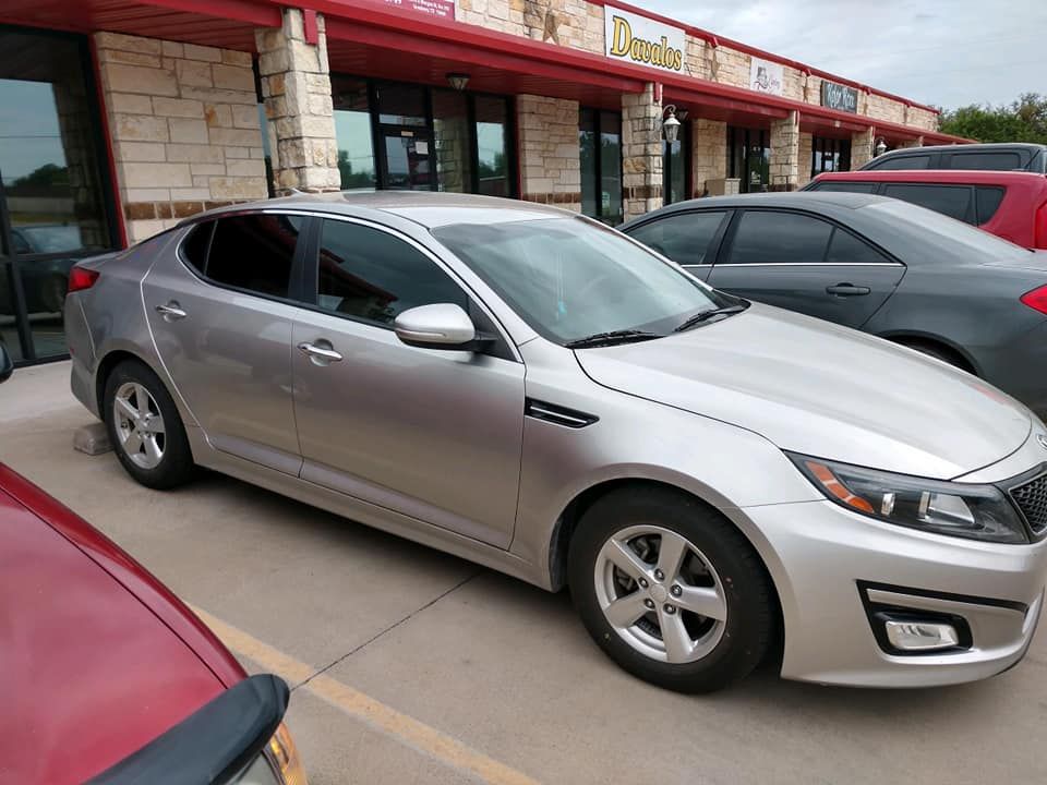 Silver Kia Optima parked in front of a strip mall with tinted windows and a red trim.
