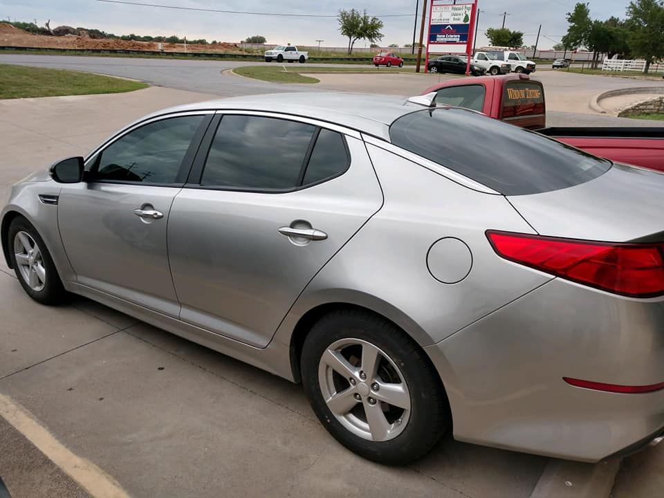 Silver Kia Optima sedan parked on pavement near a road, with tinted windows and a partially visible gas station in the background.