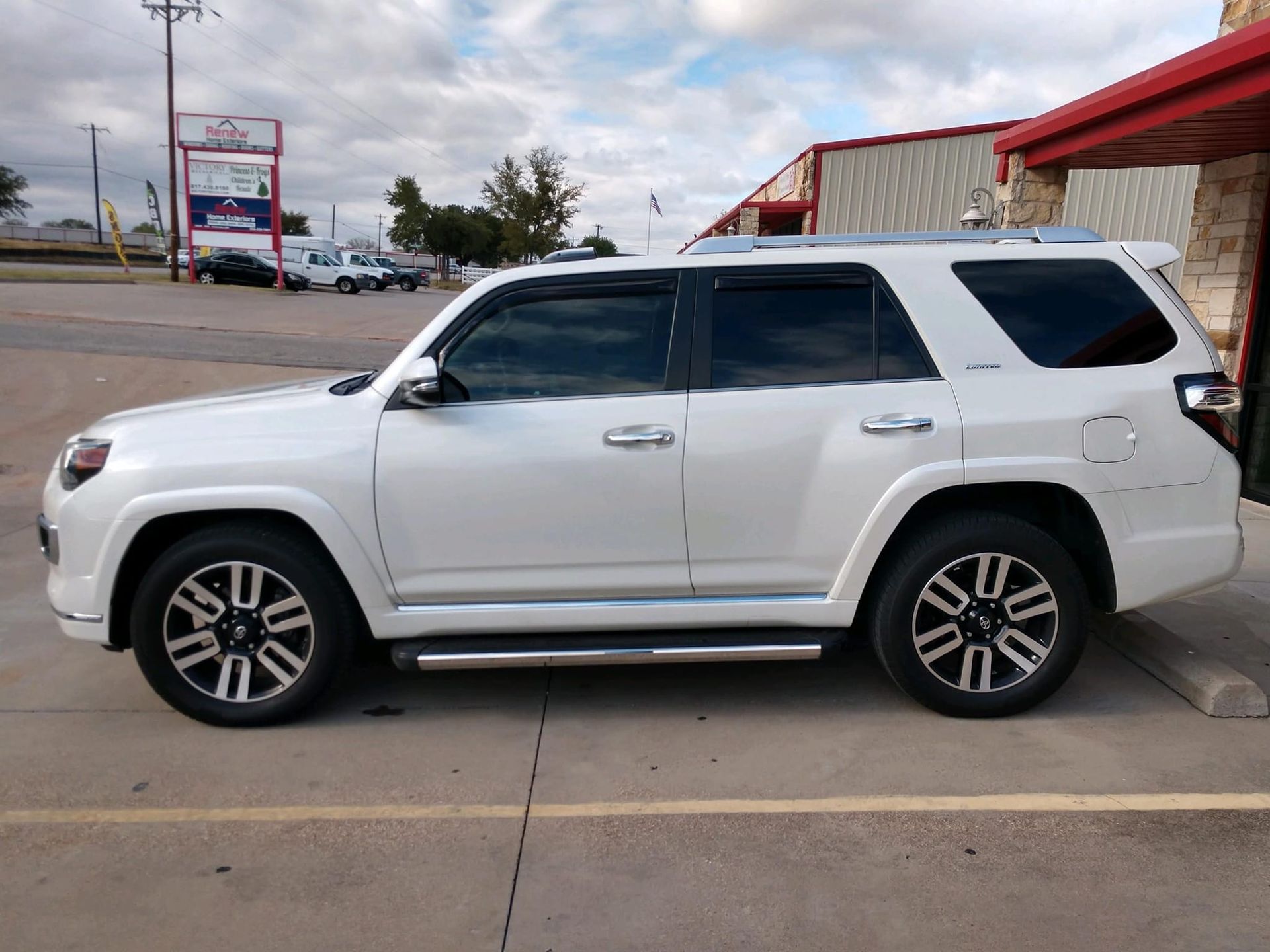 White SUV parked on pavement, chrome running boards, tinted windows, dark wheels, commercial building in background.