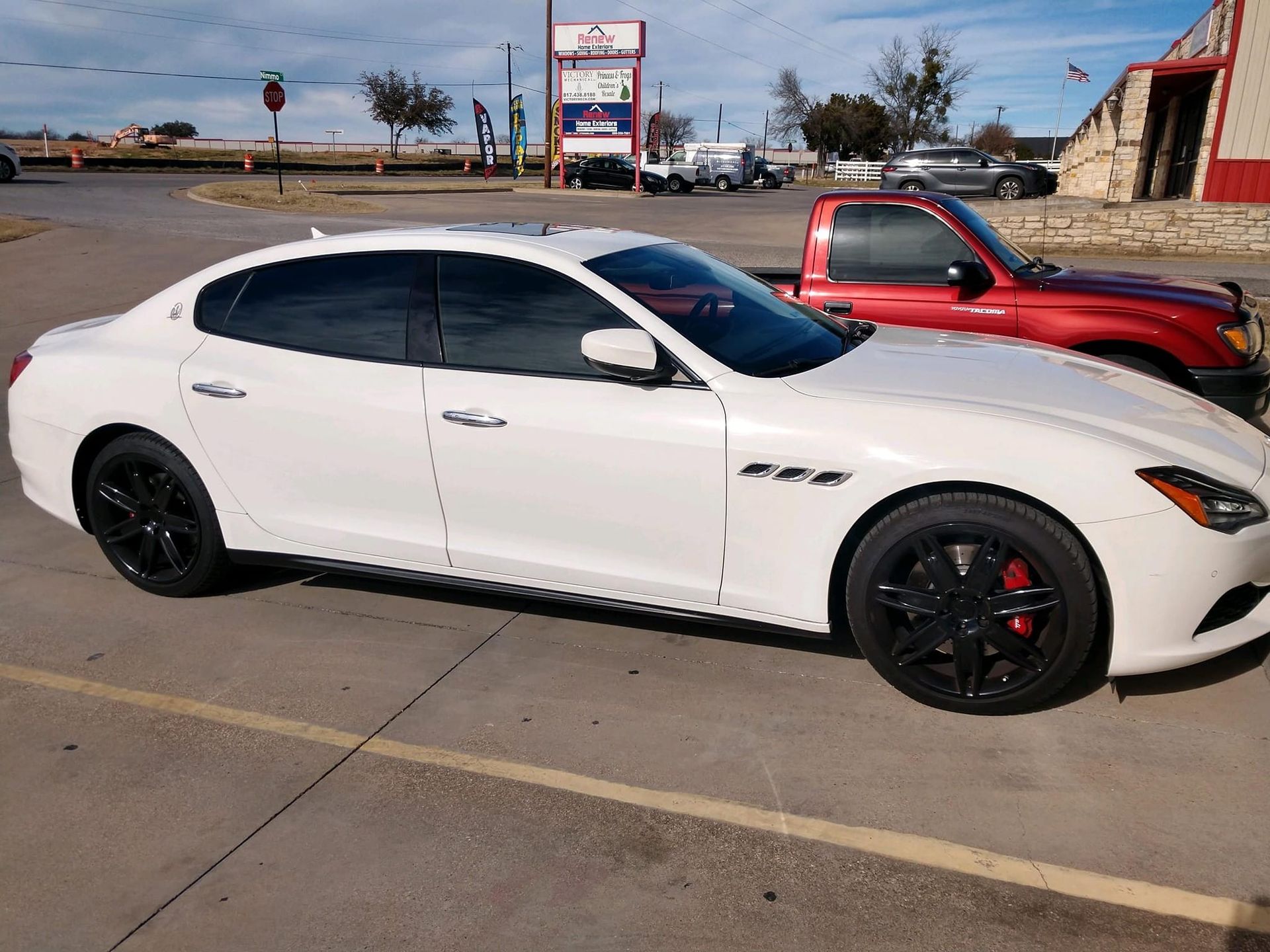 White Maserati sedan with black wheels parked next to a red truck in a parking lot.