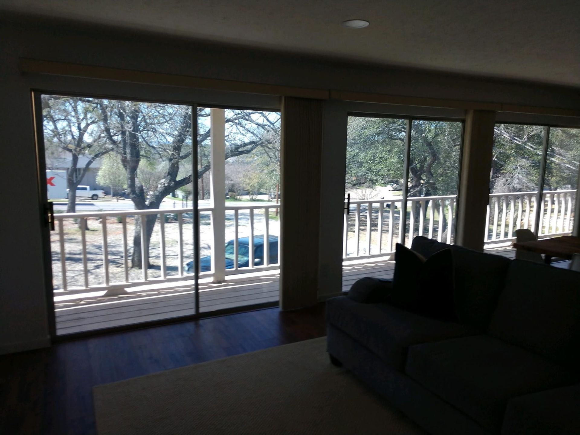 Living room with large windows overlooking a balcony and trees. Dark couch, light rug, hardwood floors.