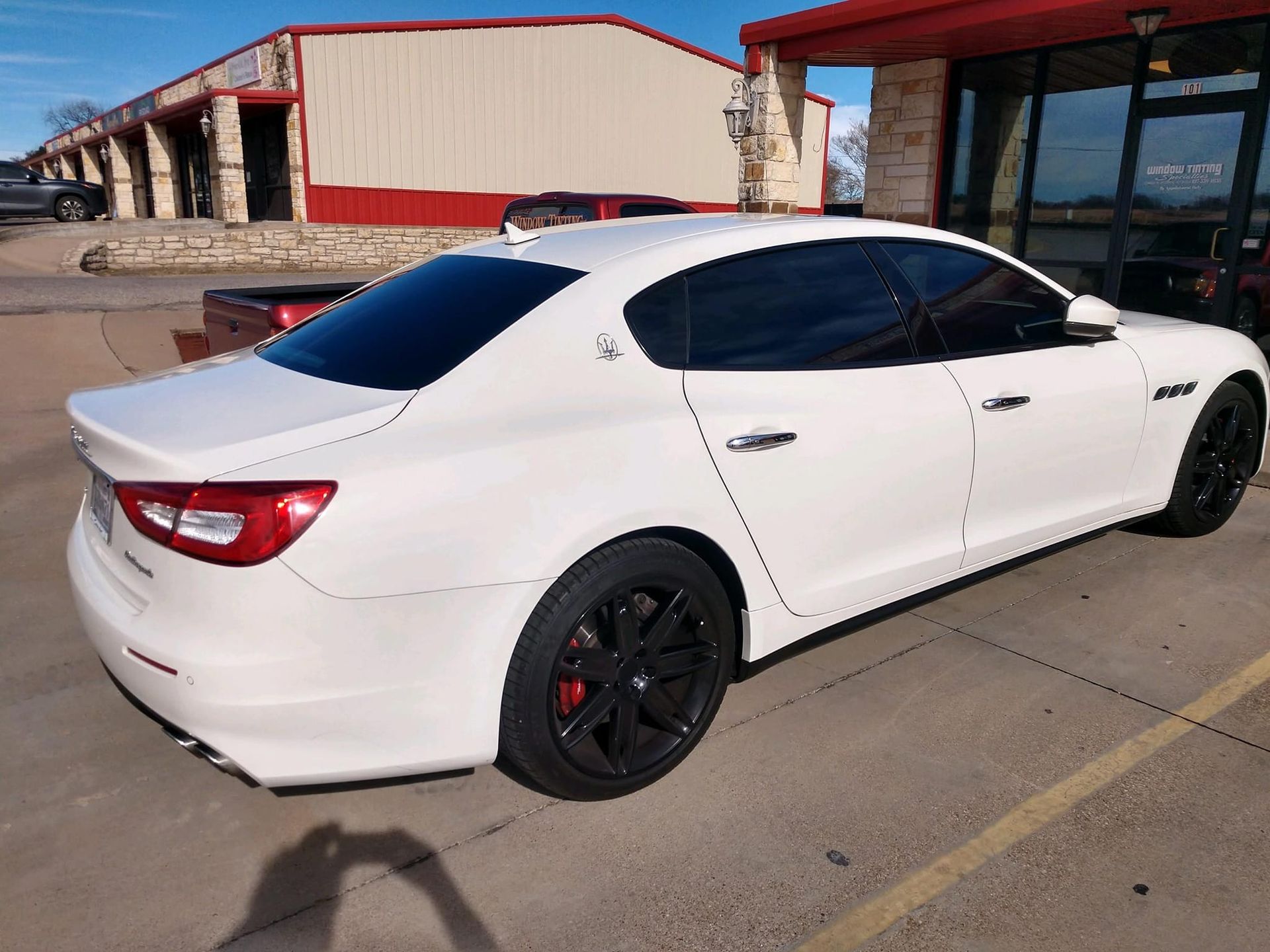 White Maserati sedan with tinted windows, black wheels, parked in front of a red-roofed building.
