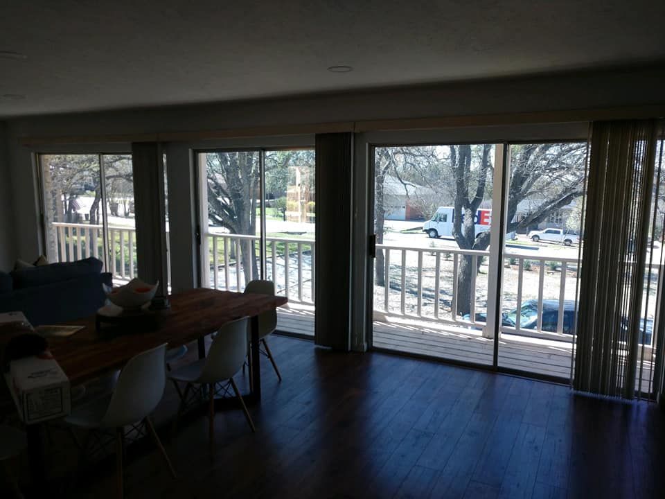 Interior view of dining area with large windows overlooking a balcony and street. Table and chairs are visible.
