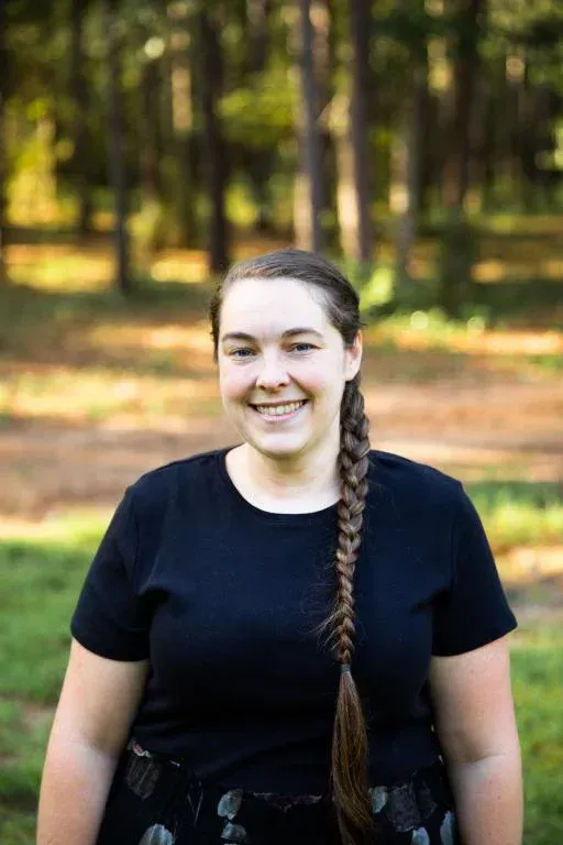 Smiling person in a black shirt standing outdoors in a wooded area with a long braid over one shoulder