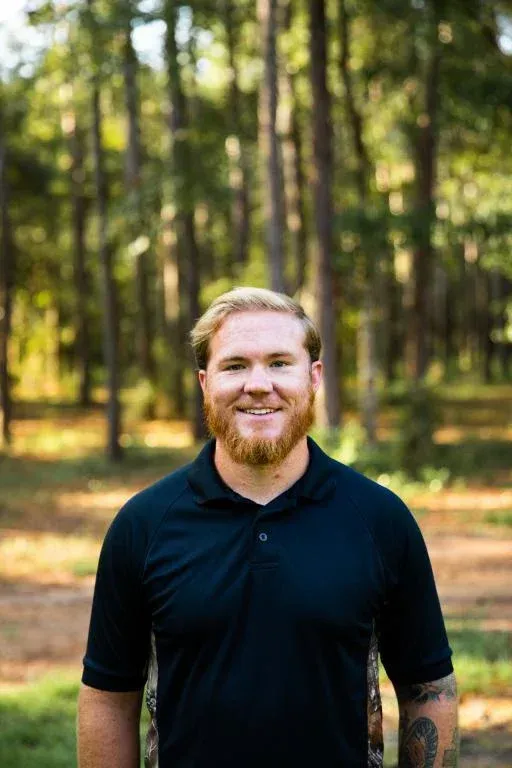 Smiling man in a black polo standing in a sunlit forest with trees in the background