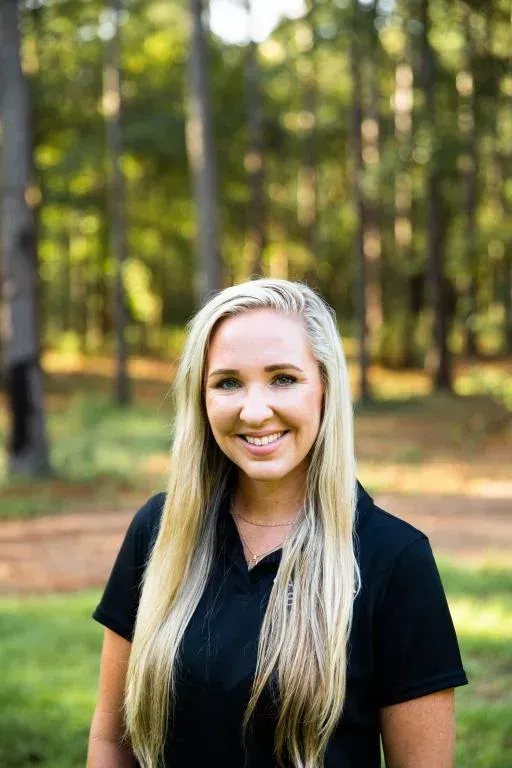 Smiling woman with long blonde hair standing outdoors in a sunlit forest clearing