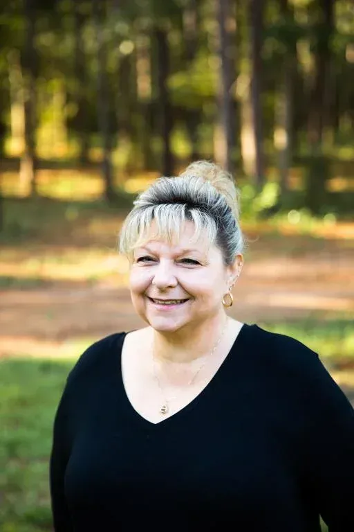 Smiling woman in a black top standing outdoors in a sunlit wooded area.