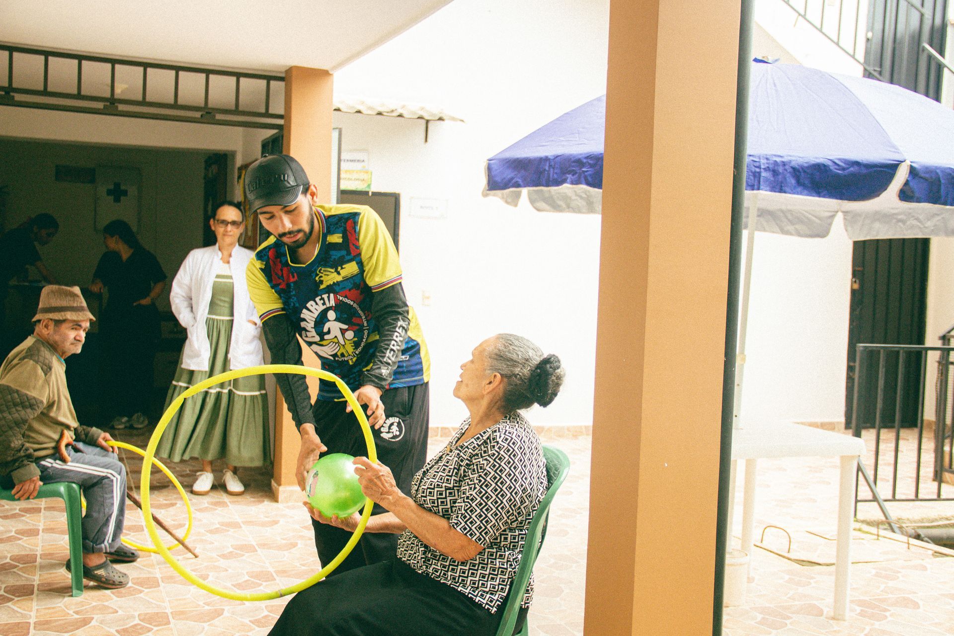 Un hombre con una camiseta colorida hace girar un aro de hula-hula para una mujer sentada en un patio; otros observan cerca.