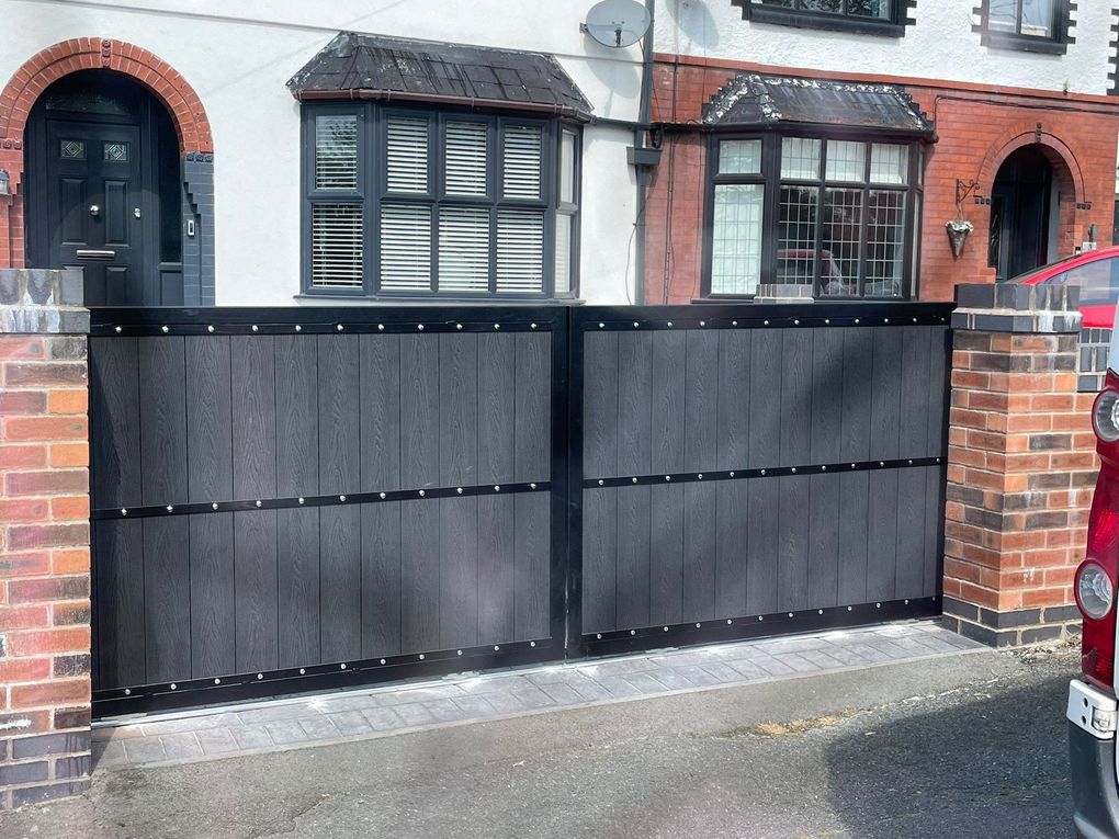 Black woodgrain composite driveway gates between brick piers in front of a bay-fronted house