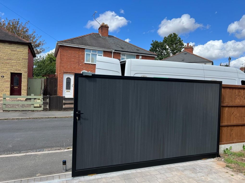Dark grey composite sliding gate in a black frame, closed across a block-paved drive