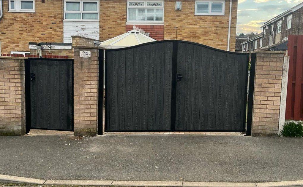Black woodgrain composite driveway gates with an arched top and a matching side gate, hung between buff brick piers