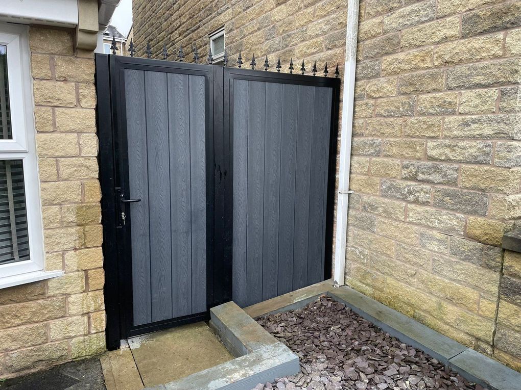 Pair of dark grey composite side gates with fleur-de-lis finials, between a stone house and a stone wall