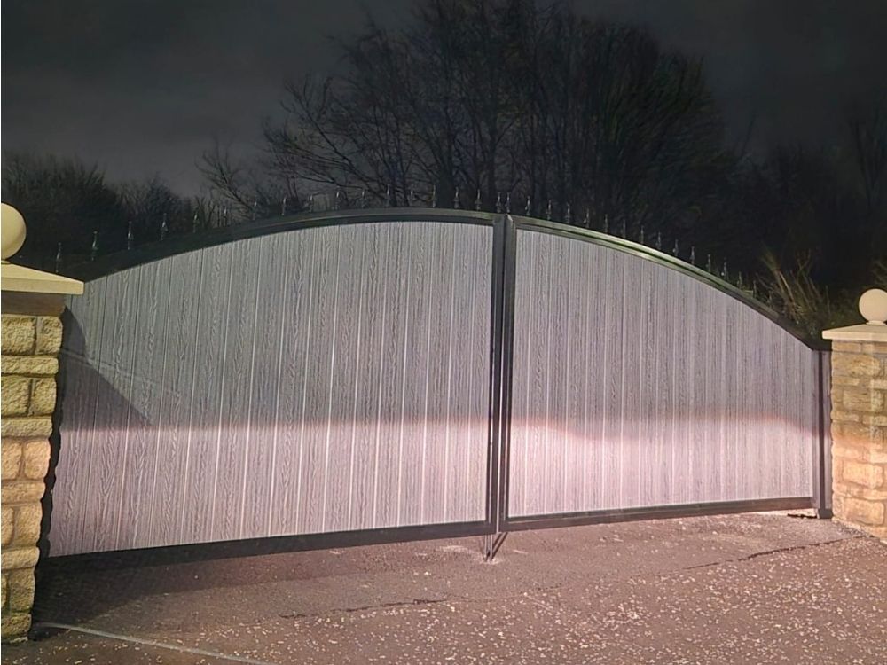 Light grey bow-top composite driveway gates with a dark frame, hung between stone-capped piers