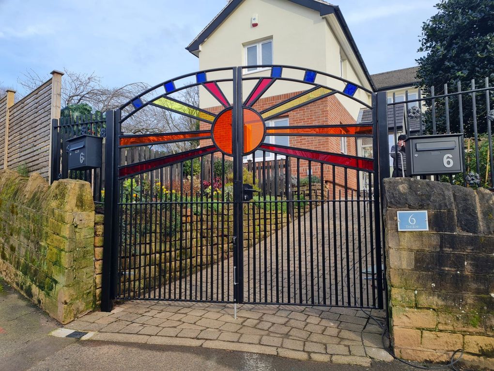 Bespoke black metal driveway gates with a bow top and a coloured acrylic sunburst infill panel in reds, oranges and blues