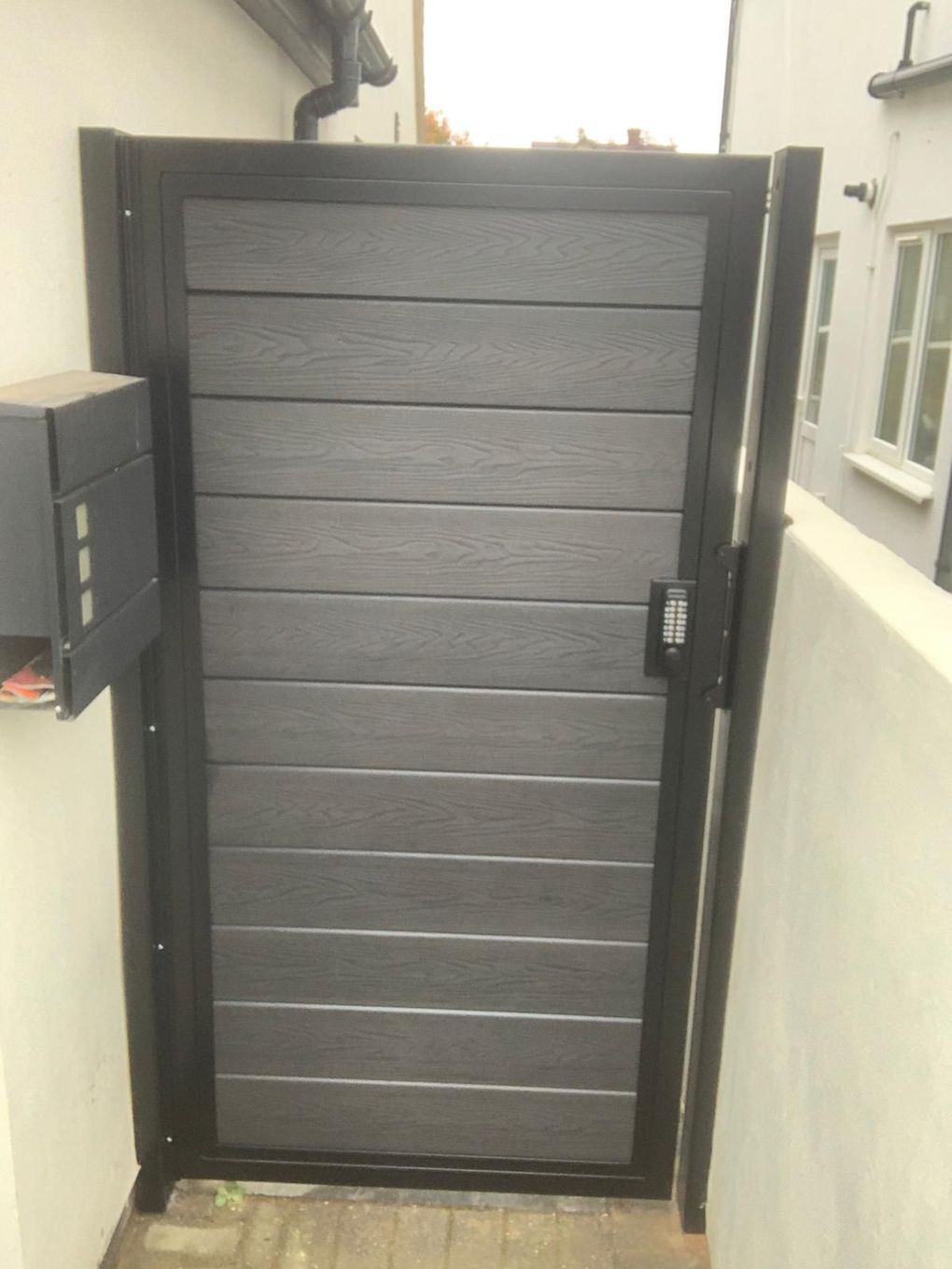 Grey composite side gate with horizontal boards and a keypad lock against a rendered wall