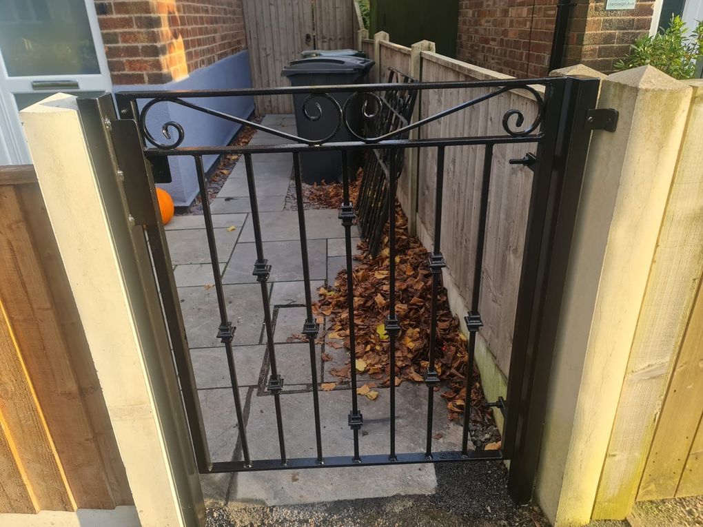 Small black metal garden gate with scroll detail and a spear finial, hung between timber fence posts