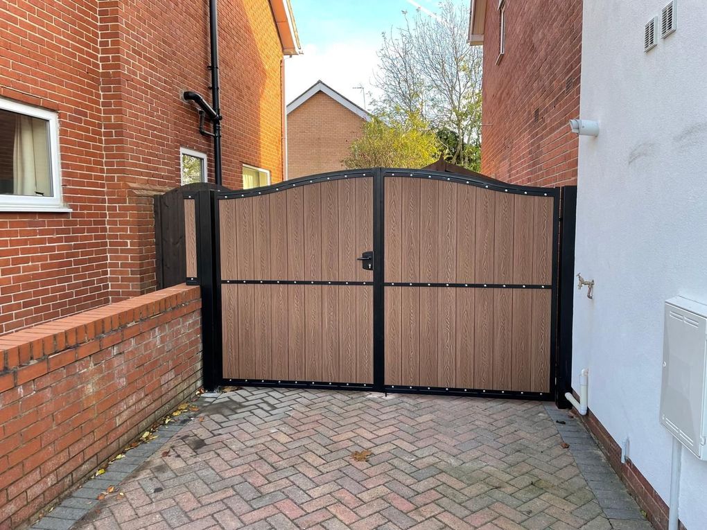 Walnut-effect composite driveway gates with an arched top and black steel frame on a block-paved drive