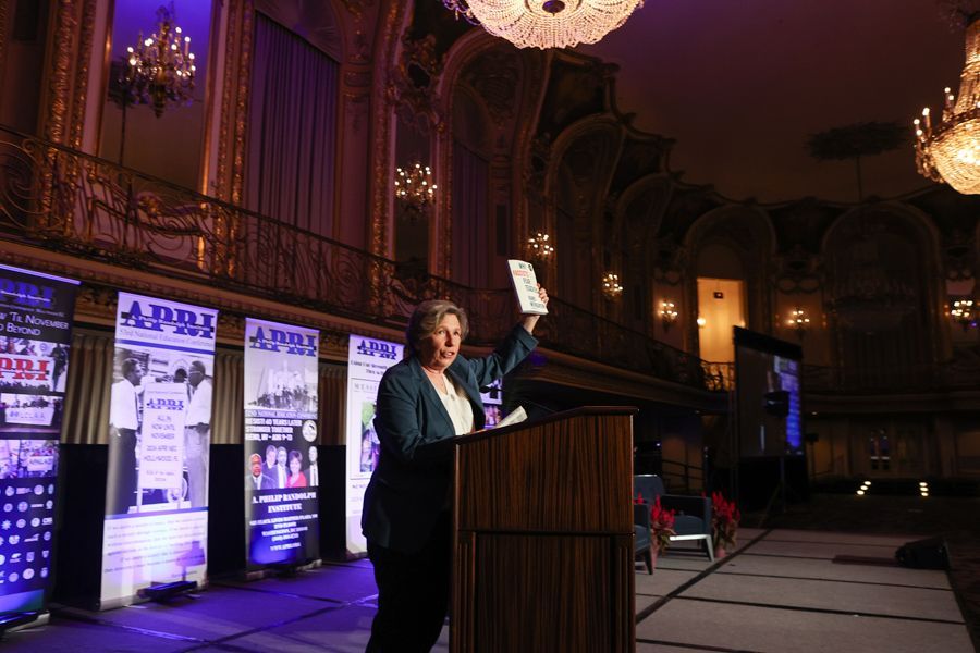 Woman at podium, holding book up, in ornate hall.
