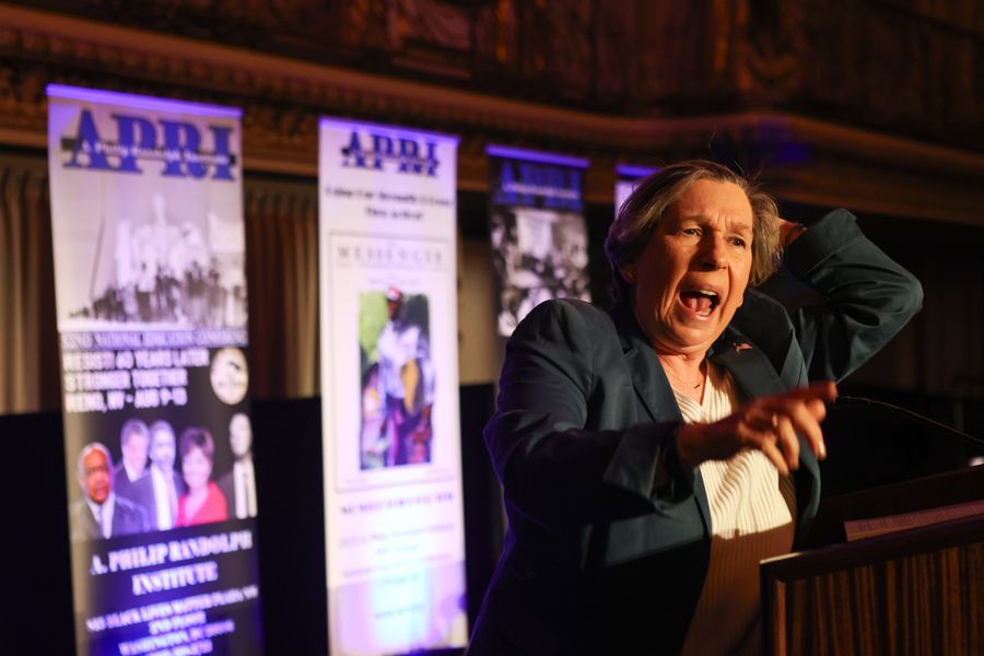 Woman speaking passionately at a podium, gesturing with hand raised. Banners with text and images are behind her.