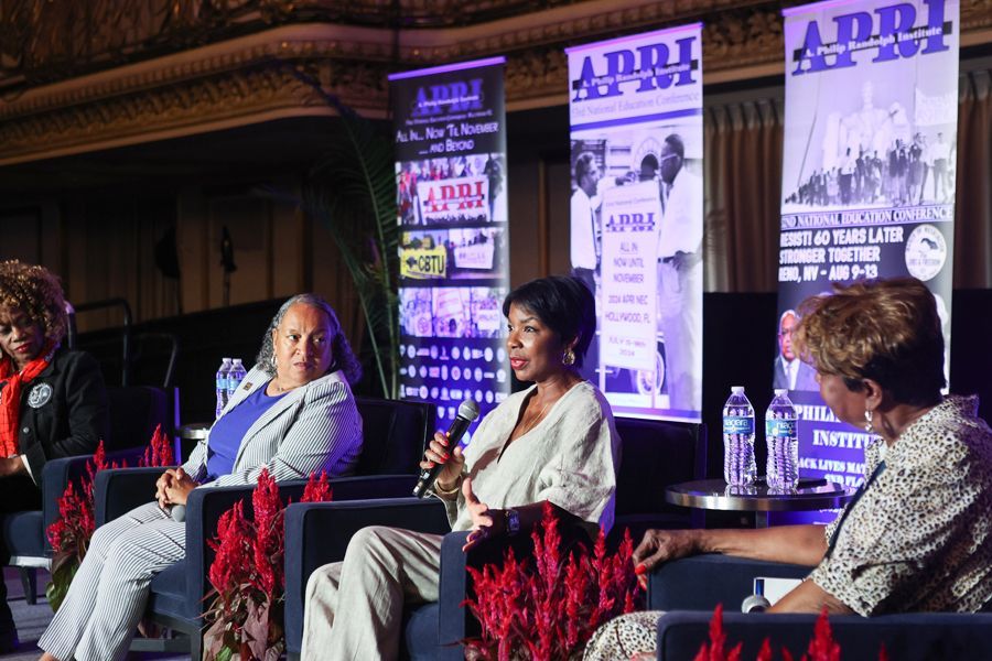 Panel of four women seated, speaking at a conference with banners in background.