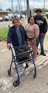 Two people standing beside an older woman using a walker on a sidewalk near a street