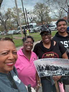 Four people smiling outdoors, holding a sign at a community event.