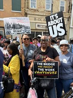 Crowd at a protest holding signs, including “It’s About Us” and “The Bridge We Cross Together.”